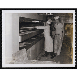 A member of the Tom Pappas Chefs' Club works with bread loaves in an industrial oven under the supervision of a kitchen staff person