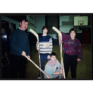 A man, two teenage boys, and a woman pose with floor hockey sticks in a gymnasium