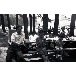 Man, teenager, and young boy sit at a picnic table in a wooded area