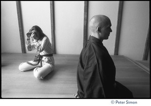 Buddhist monk seated on the floor, legs crossed, before an altar, with woman taking a photograph in the background