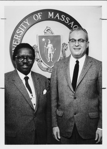 John W. Lederle standing with unidentified man in front of University of Massachusetts seal