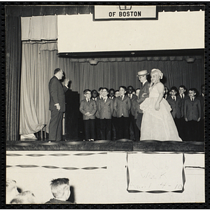 Two boys wearing costumes as a couple walk across the stage while the choir sing behind them at a Boys' Club Week celebration event. A caption on the back of the photograph states "Choral group sing about her (?) Easter bonnet"