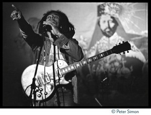 Bob Marley in concert, exhorting the audience while standing in front of a large banner with a portrait of Haile Selaisse