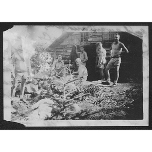 Men standing and seated around fire in front of a cabin