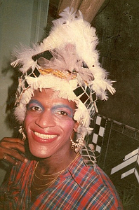 A Photograph of Marsha P. Johnson Wearing a Feather Headpiece, Blue Eye Makeup, and Plaid Shirt