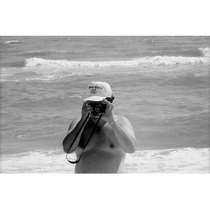 Unidentified man with a camera at the beach with the ocean behind him.