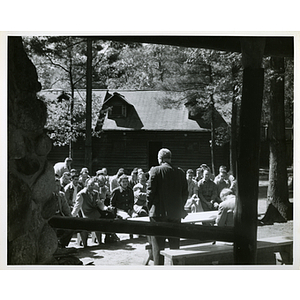 At an outdoor event, a man stands at a table while seated people look on