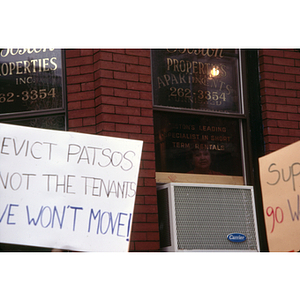Exterior view of New Boston Properties, Inc., with two protest signs in front of the building and a woman peering out the window