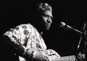 Taj Mahal in concert at Northfield, Mass.: Taj Mahal seated, playing steel guitar (blurred)