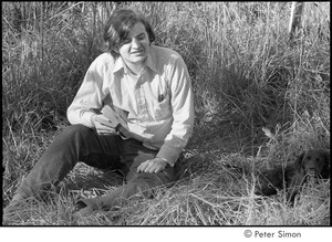 Joe Pilati in tall grass, holding a book and looking at a puppy, Packer Corners commune