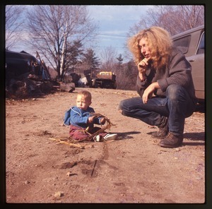 Charles and baby Eben crouched by a car (Scout) in front of the house at Montague Farm Commune