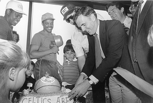 Mayor Raymond L. Flynn cutting a basketball cake with Larry Bird while Quinn Buckner, Carlos Clark as others watch on