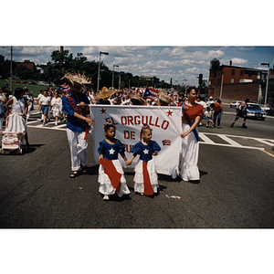 Two young girls in Puerto Rican flag dresses walk in front of a banner during the Festival Puertorriqueño parade, near Centre Street and Columbus Avenue