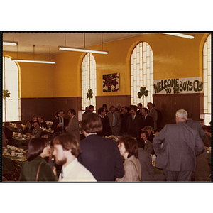 A group of men standing in front of a wall while others sit and stand around tables during a Boys' Club St. Patrick's Day event