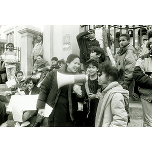 Woman hands the megaphone to a young girl to speak at a bilingual education rally from the steps of the Massachusetts State House, with adult activists and children in the background