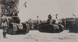 Street-level view of two tanks rolling through the Arc de Triomphe at the end of the parade; soldiers stand in the tank turrets holding French flags, Paris