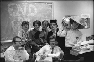 At the Boston University News Office: staff of BU News, Don McClean holding sign, Peter Simon top left, unidentified woman, unidentified man, Clif Garboden, Ed Siegel, and Joe Pilati in front with typewriter, December 1967