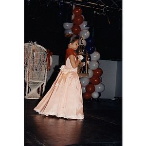 A little girl walks across a stage carrying a large trophy during the Festival Puertorriqueño