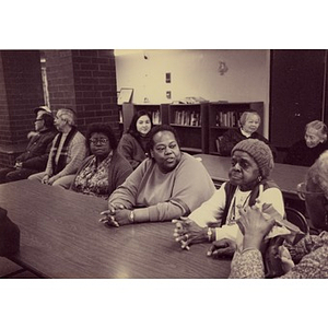 Older people seated at tables in a common area, perhaps waiting for a meeting to start.