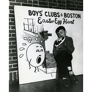 A boys strikes a humorous pose with a large chocolate egg next to an interactive Easter poster that reads "Boys' Clubs of Boston Easter Egg Hunt"