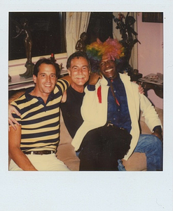 A Photograph of Marsha P. Johnson Sitting on a Friend’s Lap With Rainbow Hair and a White Jacket
