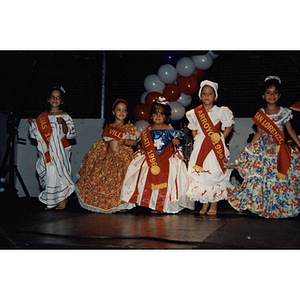 Five girls on stage in dresses and sashes at the Festival Puertorriqueño